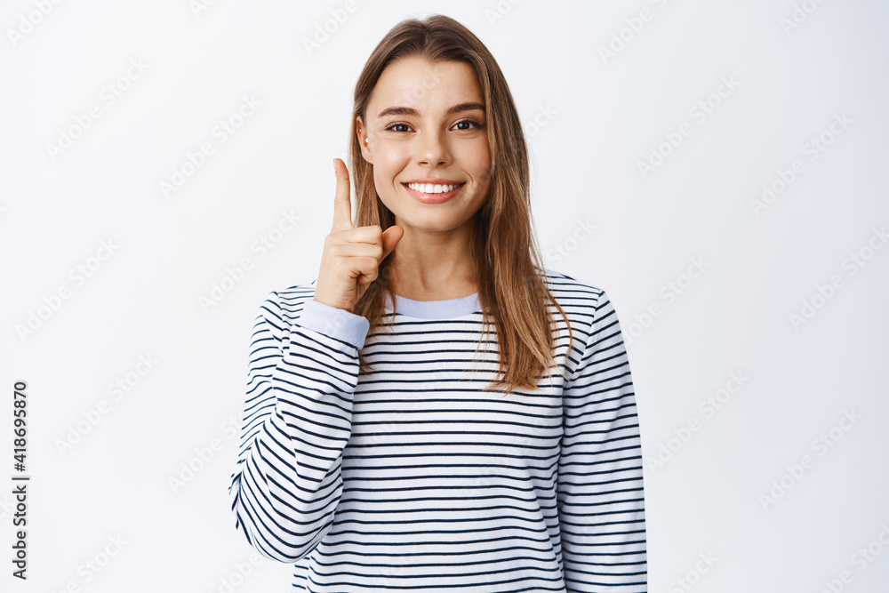 Stylish modern girl with blond hair and piercing, pointing finger up and smiling, having an idea or suggestion, wearing casual clothes, white background