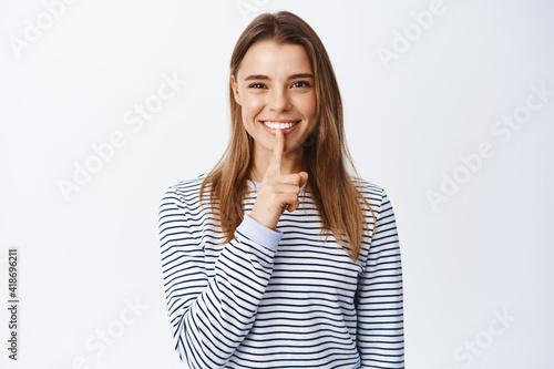 Image of pretty blond woman shushing and smiling, making shh taboo gesture and looking at camera, asking to keep quiet, sharing a secret, standing over white background