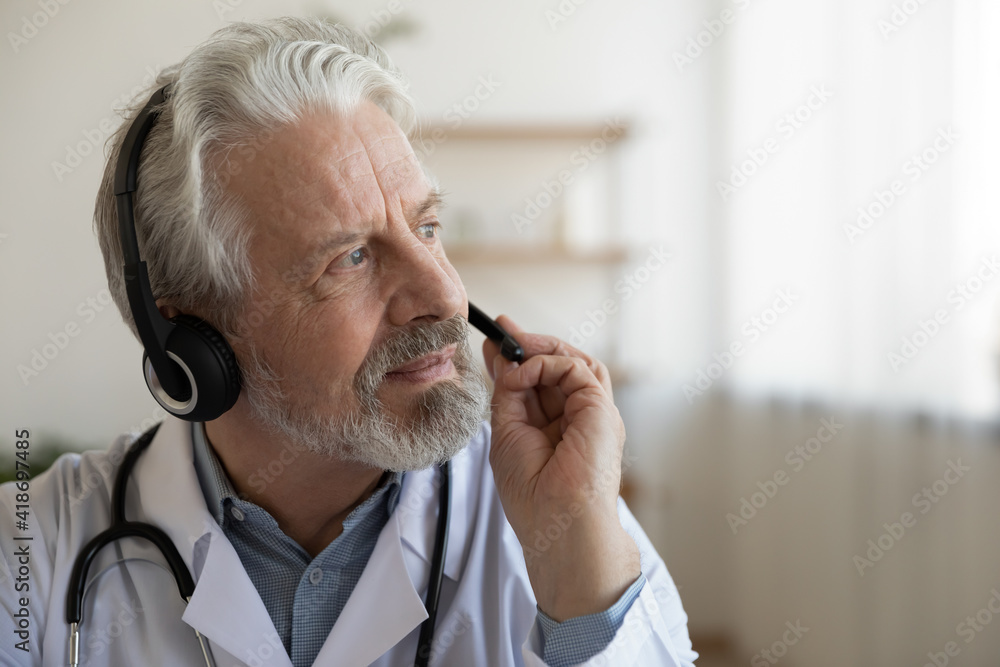Pensive mature male doctor in earphones consult patient online look in distance thinking pondering. Thoughtful senior man GP in headphones talk on video call make decision. Virtual event concept.