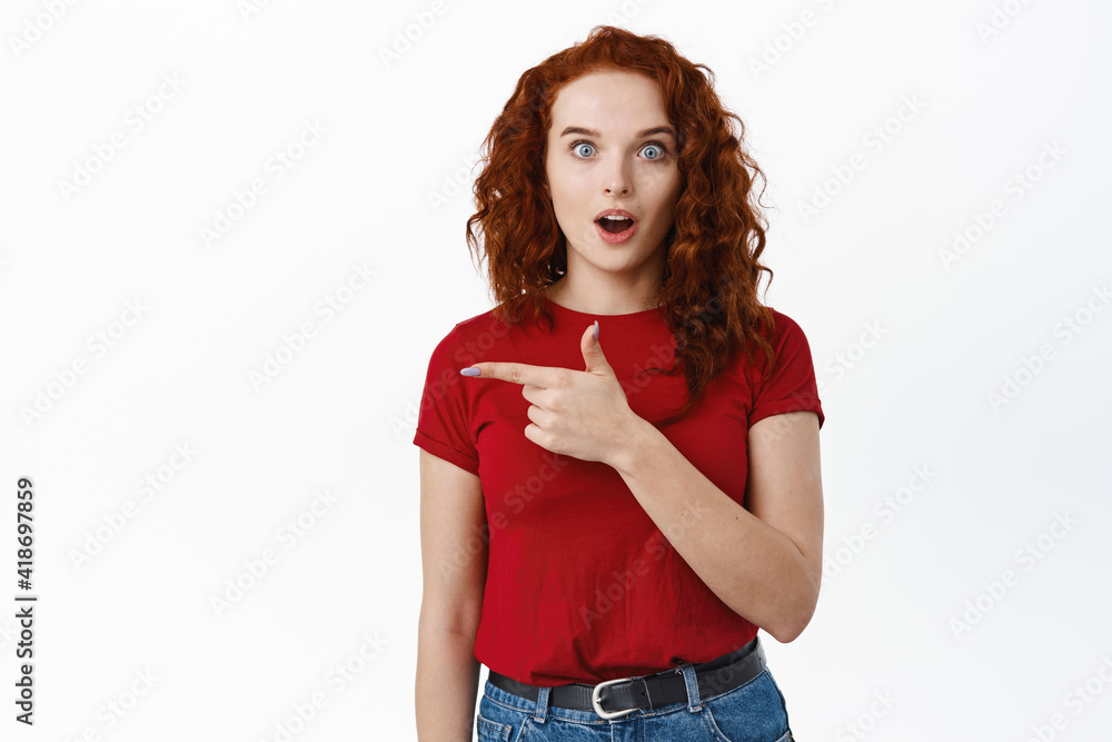 Wow this is amazing. Portrait of impressed redhead woman gasping fascinated, staring at camera while pointing finger left at banner, showing logo on white background Wow this is amazing. Portrait of impressed redhead woman gasping fascinated, staring at camera while pointing finger left at banner, showing logo on white background