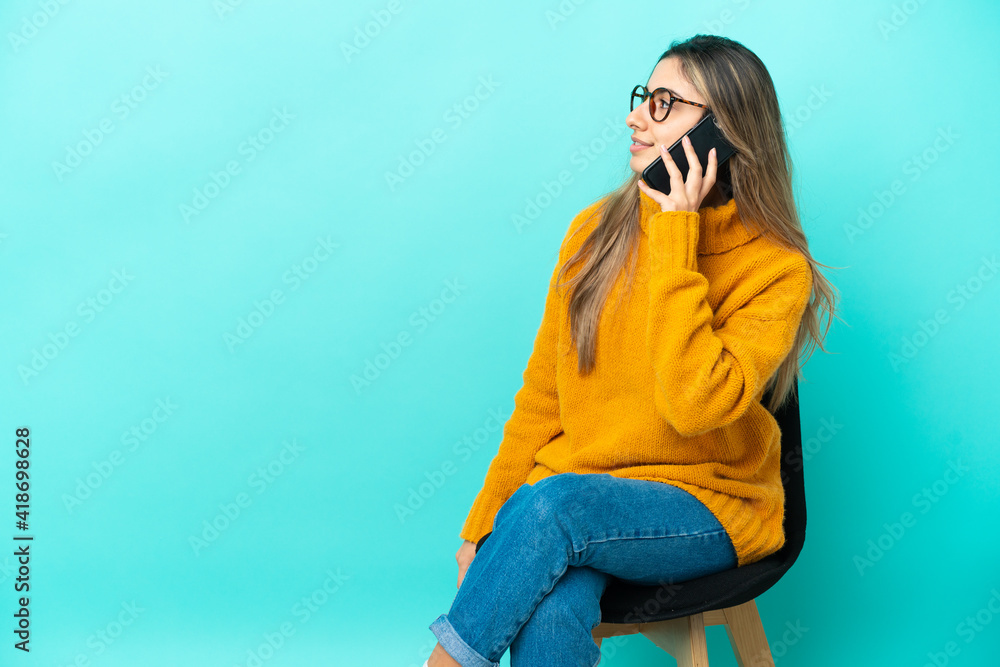 Young caucasian woman sitting on a chair isolated on blue background keeping a conversation with the mobile phone with someone Young caucasian woman sitting on a chair isolated on blue background keeping a conversation with the mobile phone with someone