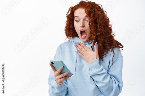 Shocked and startled redhead girl scream while reading message on smartphone, staring at mobile screen with surprised face, standing against white background