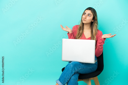 Young caucasian woman sitting on a chair with her pc isolated on blue background having doubts while raising hands © luismolinero