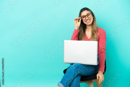 Young caucasian woman sitting on a chair with her pc isolated on blue background with glasses and happy © luismolinero
