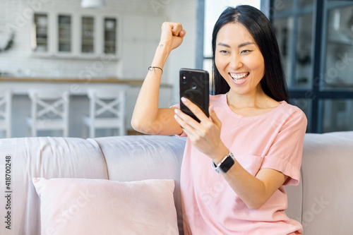 Overjoyed Asian woman holds a smartphone sitting on the sofa at home, looks at the screen and celebrates good deal or news, raised hand ecstatic, victory and triumph concept © Vadim Pastuh