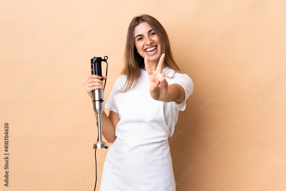 Woman using hand blender over isolated background smiling and showing victory sign Woman using hand blender over isolated background smiling and showing victory sign