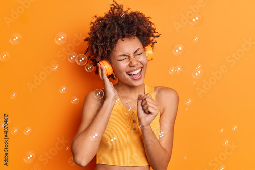 Optimistic amused Afro American woman sings song keeps hand near mouth as if microphone wears wireless headphones listens favorite music isolated over orange background with soap bubbles around photo