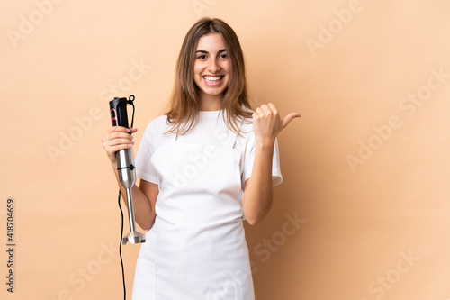 Woman using hand blender over isolated background pointing to the side to present a product