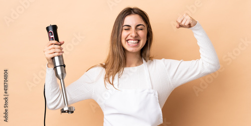 Woman using hand blender over isolated background doing strong gesture