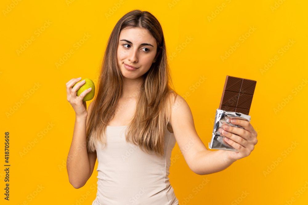 Young woman over isolated yellow background having doubts while taking a chocolate tablet in one hand and an apple in the other Young woman over isolated yellow background having doubts while taking a chocolate tablet in one hand and an apple in the other