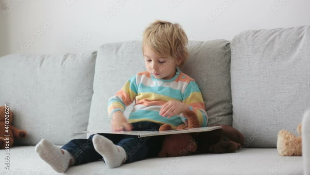 Toddler blond child, reading a book at home on the sofa with teddy bear friend Toddler blond child, reading a book at home on the sofa with teddy bear friend