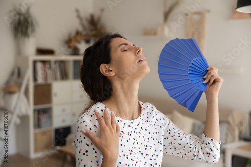 What a heat. Exhausted young female housewife suffer from extra hot temperature at home apartment wave herself use hand weaver. Overheated millennial woman cool air with paper fan miss air conditioner