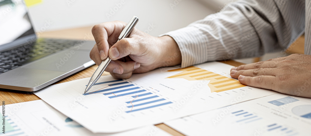 Close-up businessman holding a pen and pointing at a bar chart on a company financial document, he is analyzing historical financial data to plan how to grow the company. Financial concept. Close-up businessman holding a pen and pointing at a bar chart on a company financial document, he is analyzing historical financial data to plan how to grow the company. Financial concept.