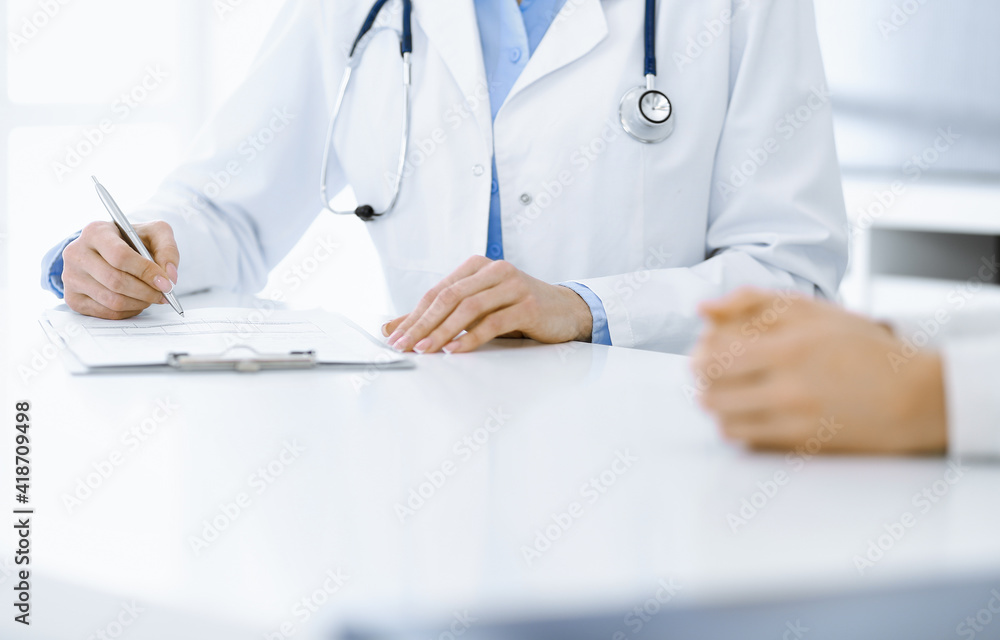 Unknown woman-doctor and female patient sitting and talking at medical examination in clinic, close-up. Therapist wearing blue blouse is filling up medication history record. Medicine concept Unknown woman-doctor and female patient sitting and talking at medical examination in clinic, close-up. Therapist wearing blue blouse is filling up medication history record. Medicine concept