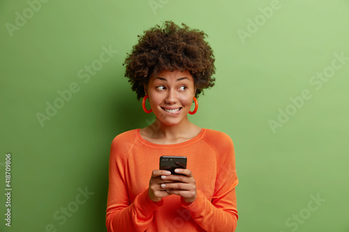 Pensive dark skinned young woman bites lips holds modern cellular uses mobile phoe sends text messages wears orange jumper and earrings isolated over green background. People technology concept photo