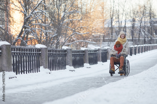 Wide angle view at African American man using wheelchair having fun outdoors in winter with smiling young woman assisting, copy space