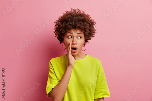 Shocked mysterious curly haired woman keeps hand near opened mouth tells secret spreads rumors whispers something secretly concentrated aside wears casual t shirt isolated over pink background