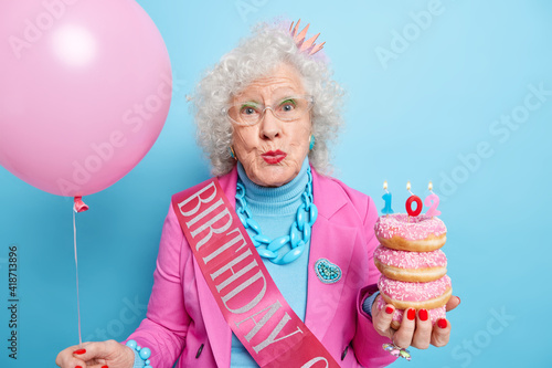 Beautiful curly haired senior woman keeps lips folded enjoys birthday celebration holds pile of delicious doughnuts with candles inflated balloon isolated over blue background. Celebration concept