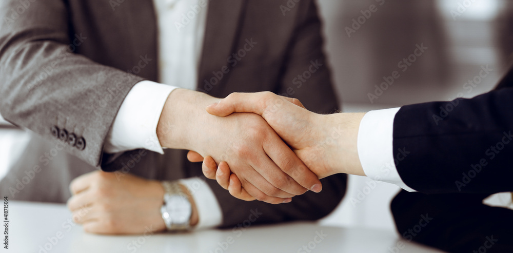 Unknown businessman shaking hands with his colleague or partner above the glass desk in modern office, close-up. Business people group at meeting