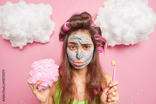 Fototapeta Naklejka Na Ścianę i Meble -  Photo of sad young brunette woman engaged in cleaning teeth facial treatments holds bath sponge and toothbrush isolated over pink background with white clouds above. Stomatology beauty concept