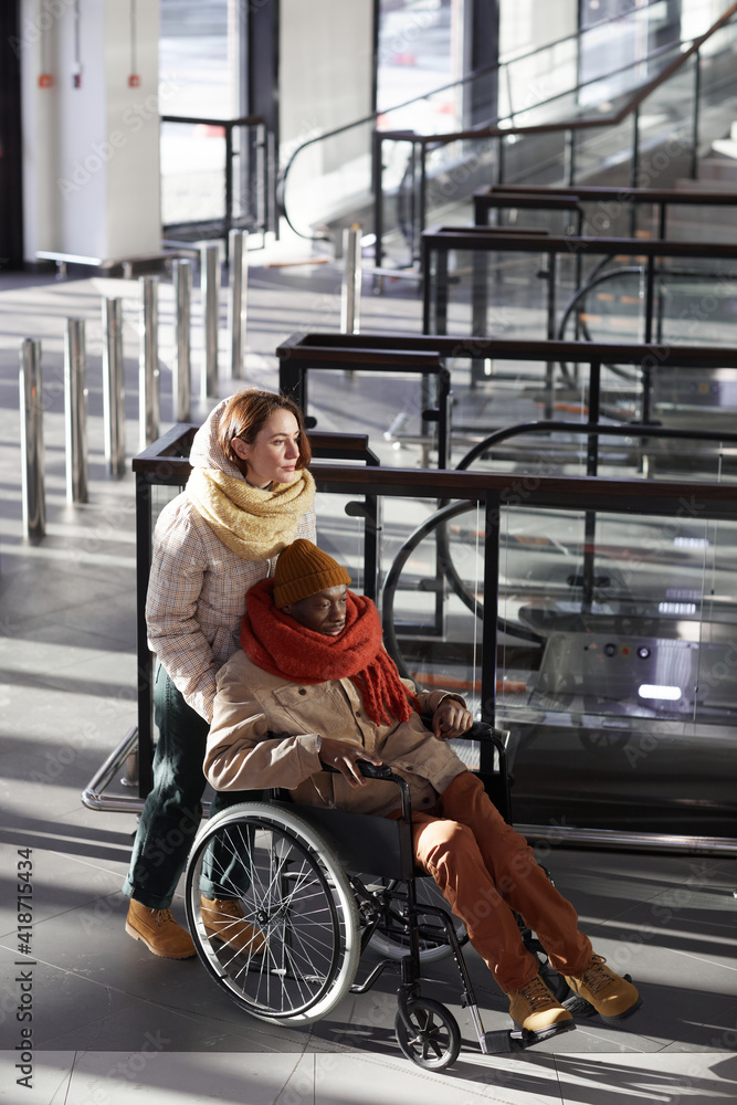 Obraz premium Vertical full length portrait of young woman assisting African American man in wheelchair at subway station, copy space