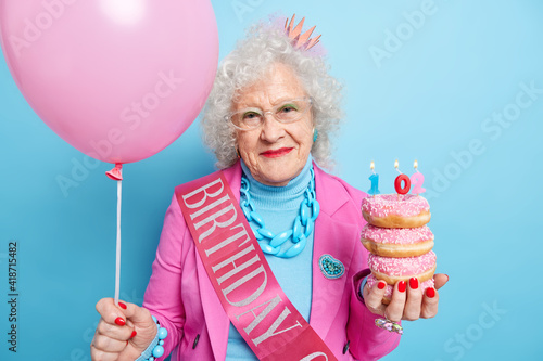 People age holidays festive event concept. Beautiful old woman with curly hair wrinkled face holds glazed doughnuts inflated balloon celebrates bithday infamily circle isolatd over blue background