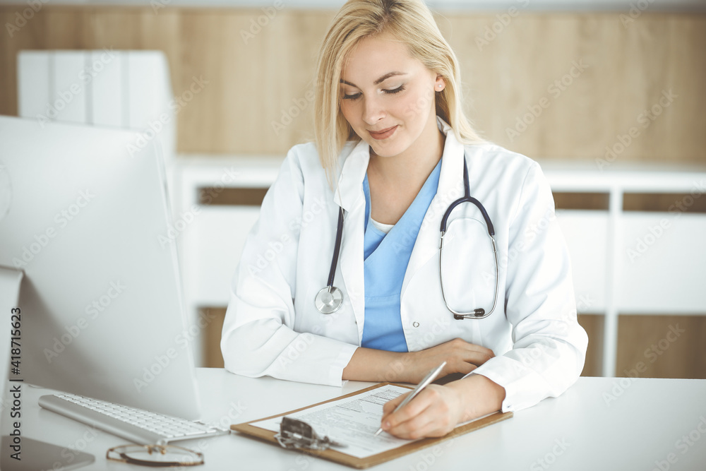 Woman-doctor at work while sitting at the desk in hospital or clinic. Blonde cheerful physician filling up medication history record form Woman-doctor at work while sitting at the desk in hospital or clinic. Blonde cheerful physician filling up medication history record form