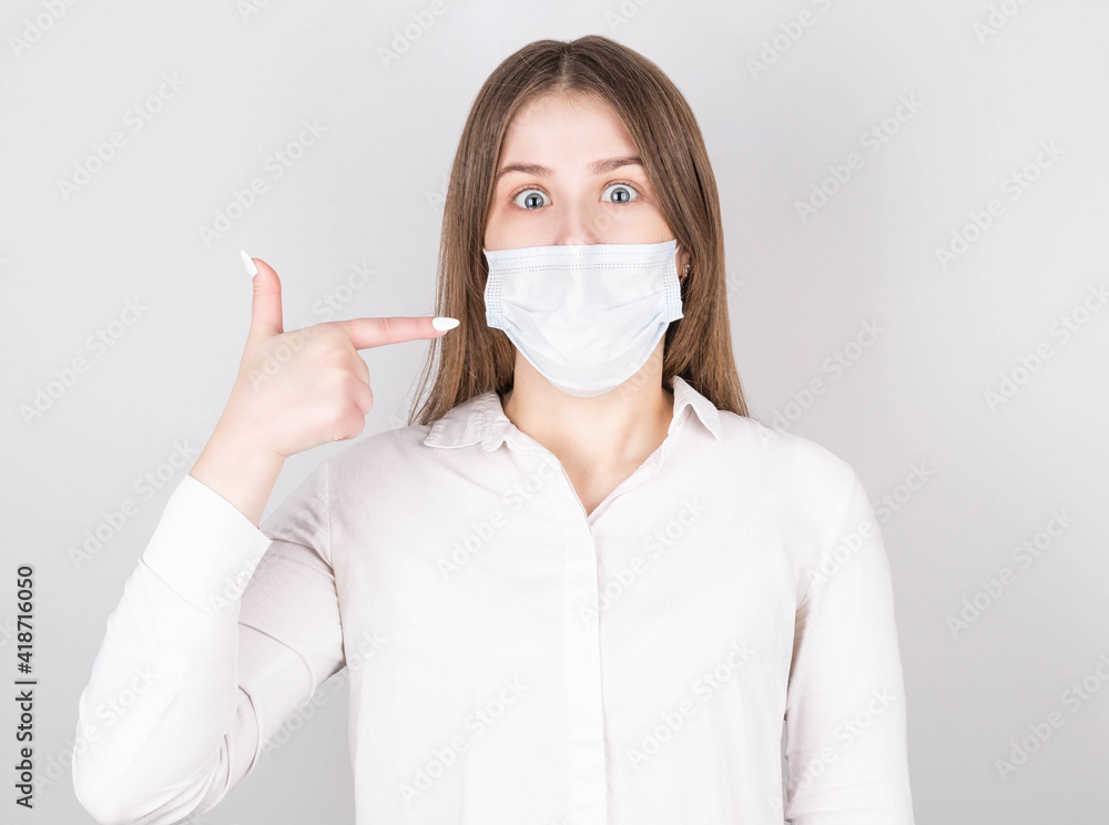 Portrait of surprised girl in medical mask and blouse points to new covid-29 safety clothing, dressed in casual style, isolated on gray colored background Portrait of surprised girl in medical mask and blouse points to new covid-29 safety clothing, dressed in casual style, isolated on gray colored background