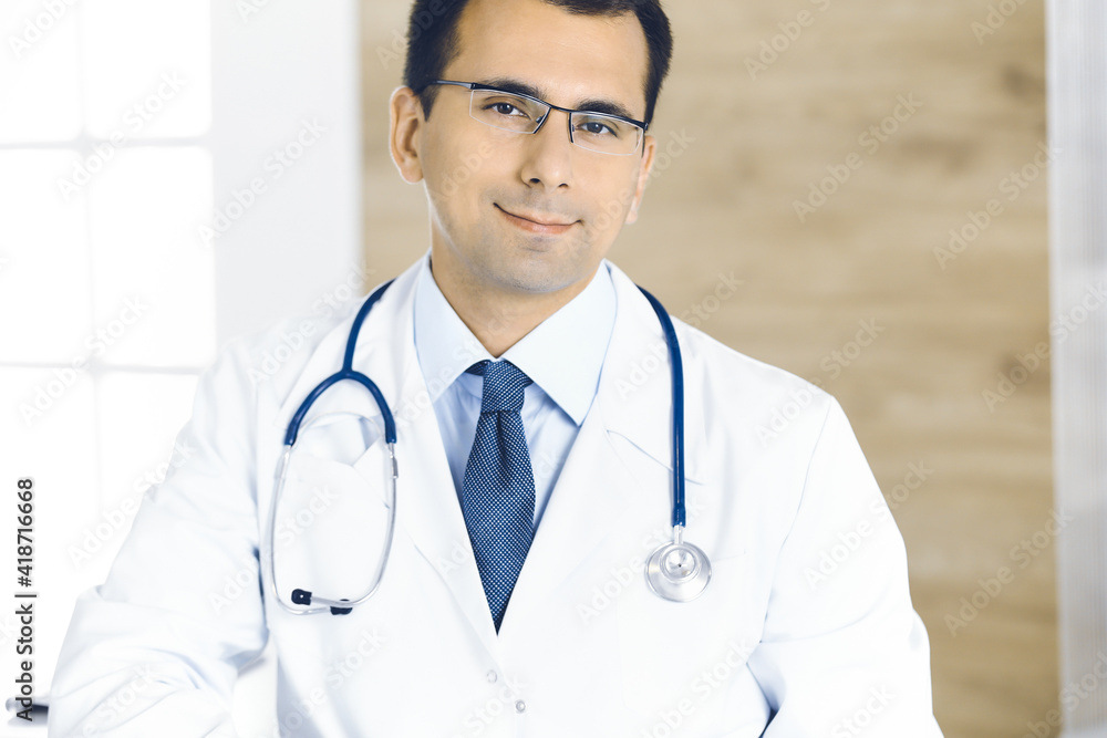 Man-doctor sitting at the desk at his working place and smiling at camera. Perfect medical service in clinic Man-doctor sitting at the desk at his working place and smiling at camera. Perfect medical service in clinic