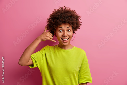 Call me sometimes. Positive dark skinned young woman makes phone gesture near ear wants to have conversation with you speaks to person dressed in casual t shirt poses against pink background