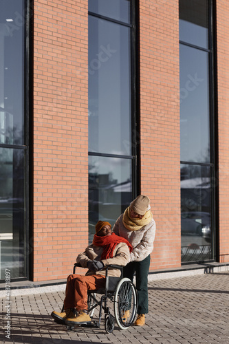 Vertical full length view at African American man in wheelchair moving in urban city with young woman assisting, copy space