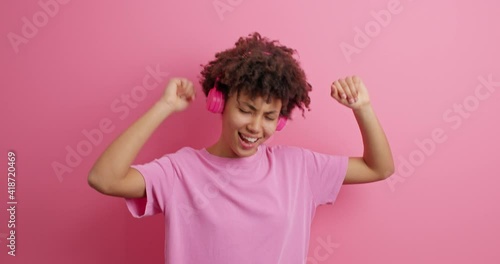 Joyful carefree Afro American girl carried away with music enjoys favorite playlist in wireless headphones has fun moves with thythm of song wears casual t shirt isolated over pink background photo