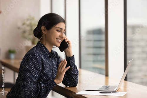 Close up smiling Indian woman talking, holding smartphone, using laptop, manager businesswoman consulting client by phone call, looking at computer screen, discussing project, reading information