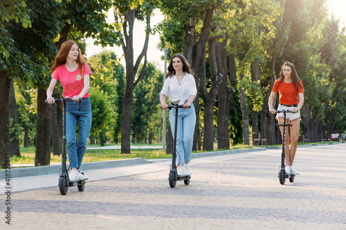 Three young girl friends on the electro scooters having fun in city street at summer sunny day. Outdoor portrait of three friends girl riding electric kick scooter in the park