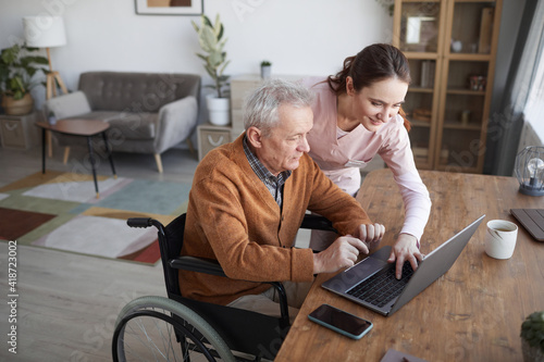 Portrait of senior man in wheelchair using laptop at retirement home with nurse assisting him, copy space photo