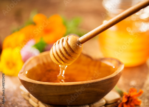 Honey dripping from honey dipper in wooden bowl.  Close-up. Healthy organic Thick honey dipping from the wooden honey spoon, closeup. Flowers and jar on the table