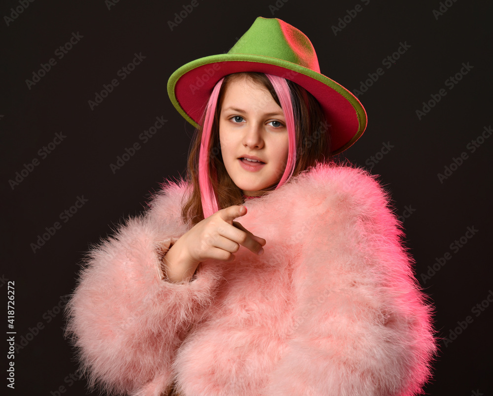 fashionable girl in bright clothes looks to the side and puts a gun from her fingers. A girl in a fluffy coat, hat and with strands in her hair folded her fingers with a gun, on a black background fashionable girl in bright clothes looks to the side and puts a gun from her fingers. A girl in a fluffy coat, hat and with strands in her hair folded her fingers with a gun, on a black background