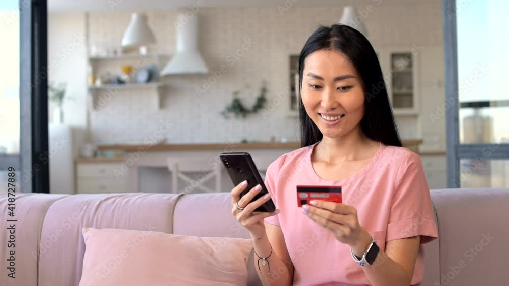 Cheerful young Asian woman making an order online, holds a smartphone and a debit cad, pays for the purchase by the phone, a multiracial girl is using mobile phone for online shopping Cheerful young Asian woman making an order online, holds a smartphone and a debit cad, pays for the purchase by the phone, a multiracial girl is using mobile phone for online shopping