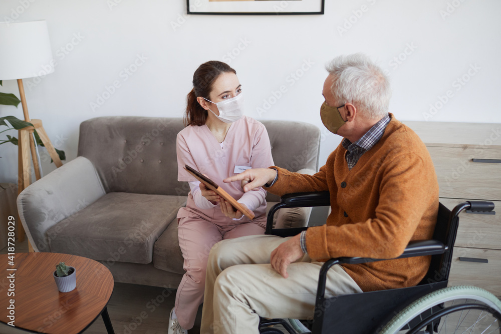 High angle portrait of young female nurse assisting senior man in wheelchair using digital tablet at retirement home, both wearing masks, copy space High angle portrait of young female nurse assisting senior man in wheelchair using digital tablet at retirement home, both wearing masks, copy space