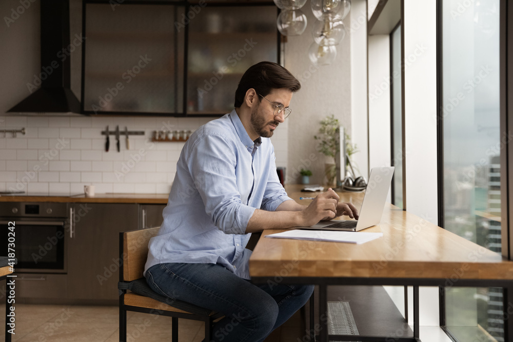 Side view confident businessman wearing glasses using laptop at home, looking at screen, sitting in kitchen, browsing apps, working on online project, accounting, student searching information Side view confident businessman wearing glasses using laptop at home, looking at screen, sitting in kitchen, browsing apps, working on online project, accounting, student searching information
