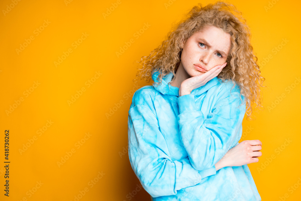 Adorable sad girl with blond hair in a blue sweater leans on her arm while standing on an orange background Adorable sad girl with blond hair in a blue sweater leans on her arm while standing on an orange background