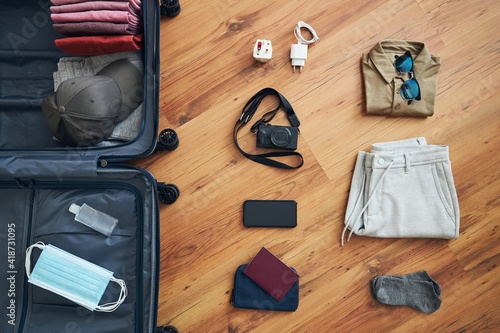 Preparing for travel in new normal. Packing suitcase with summer clothes, smart phone, camera, passport and objects for personal protection - face mask and hand sanitizer.
