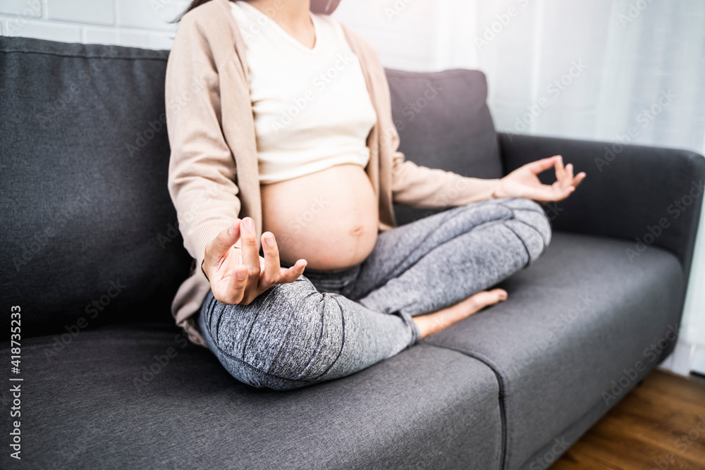 close up asian pregnant woman meditating to cope with stress, ache and pain on pregnancy hormones, meditation sitting on sofa hand on knee position, with white brick wall background and a plant pot