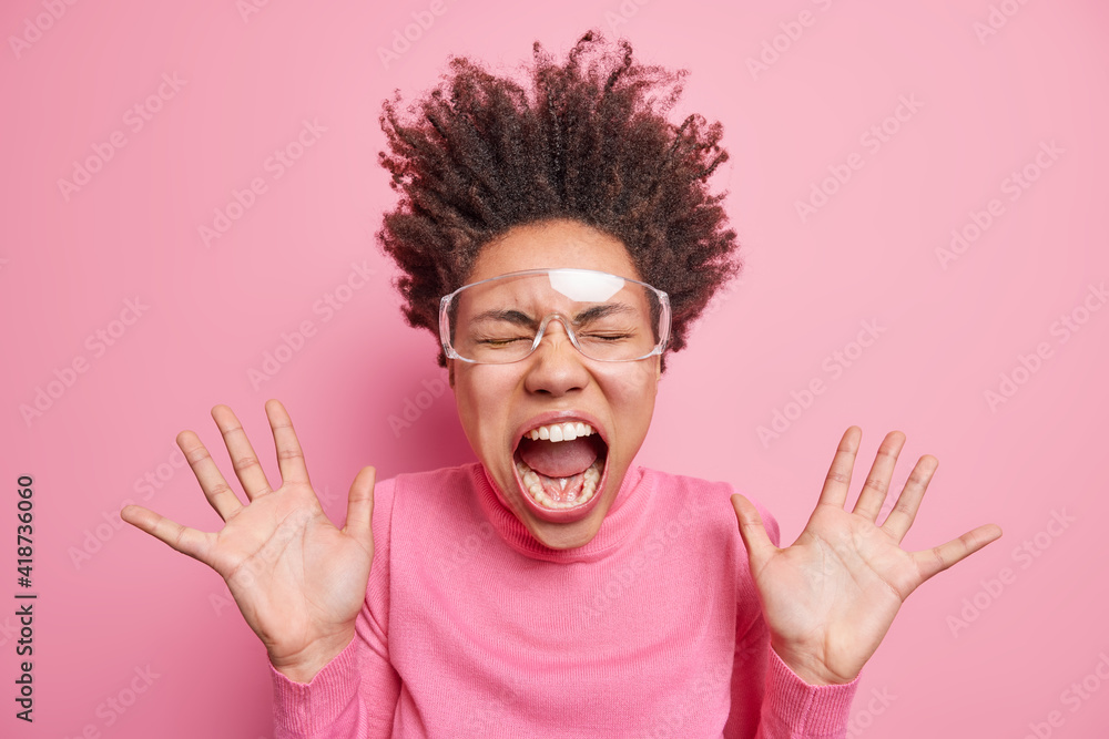 Super excited emotional Afro American woman goes carazy and screams with wide opened mouth keeps palms raised has curly hair standing on end wears transparent glasses and pink jumper. Emotions concept Super excited emotional Afro American woman goes carazy and screams with wide opened mouth keeps palms raised has curly hair standing on end wears transparent glasses and pink jumper. Emotions concept