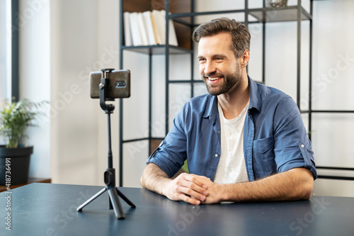 Smiling mature handsome man blogger, stylish, sitting in front of cellphone on tripod stabilizer recording streaming video, sharing shopping experience, recommending or evaluating service online