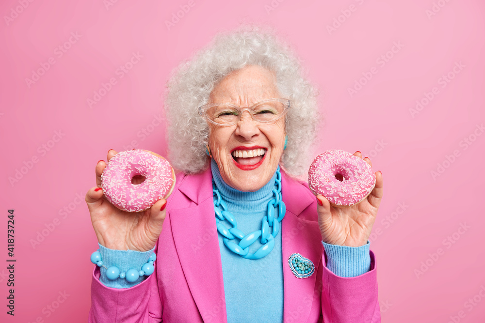 Charming happy mature woman smiles broadly shows white perfect teeth holds two glazed doughnuts dressed in fashionable festive clothes with jewelry isolated over pink background. Old age concept