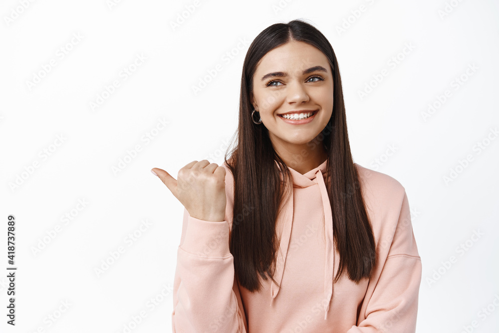 Portrait of smiling brunette female model in hoodie, pointing and looking left at advertisement, happy gazing at product, demonstrating logo, white background Portrait of smiling brunette female model in hoodie, pointing and looking left at advertisement, happy gazing at product, demonstrating logo, white background