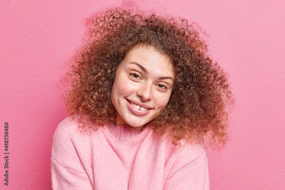Portrait of positive alluring woman with curly bushy hair smiles gently at camera shows white teeth has healthy skin wears casual jumper isolated over pink background. Natural beauty concept Portrait of positive alluring woman with curly bushy hair smiles gently at camera shows white teeth has healthy skin wears casual jumper isolated over pink background. Natural beauty concept