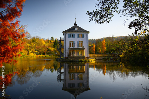 A baroque house, called Trappenseeschlösschen, surrounded by Lake Trappensee in Heilbronn, Germany during autumn. The house and the colorful trees reflect in the water.