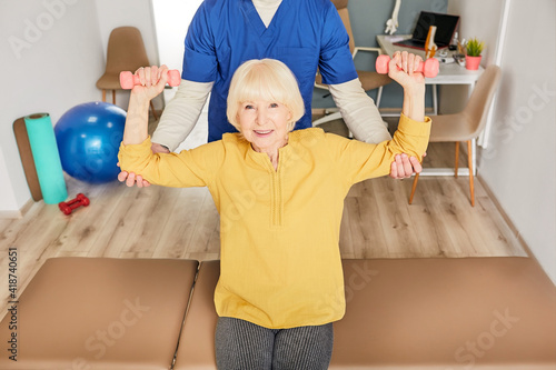Physiotherapist helping a senior woman to lift dumbbells for rehabilitation after injury. Physio treatment at a rehab center